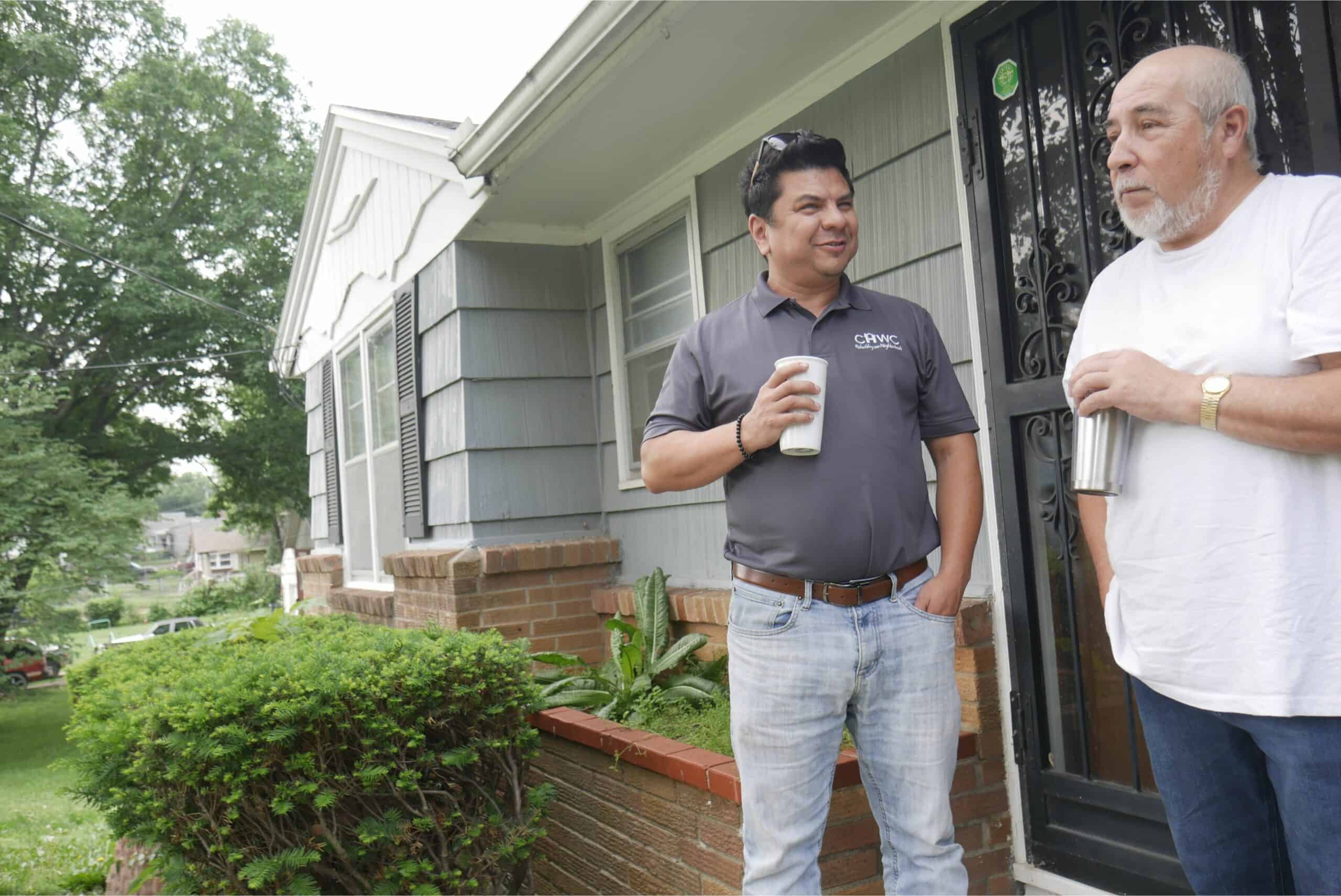 Lisandro, a housing counselor at CHWC, and David, a KCK homeowner, stand on David's front porch talking and drinking coffee. 