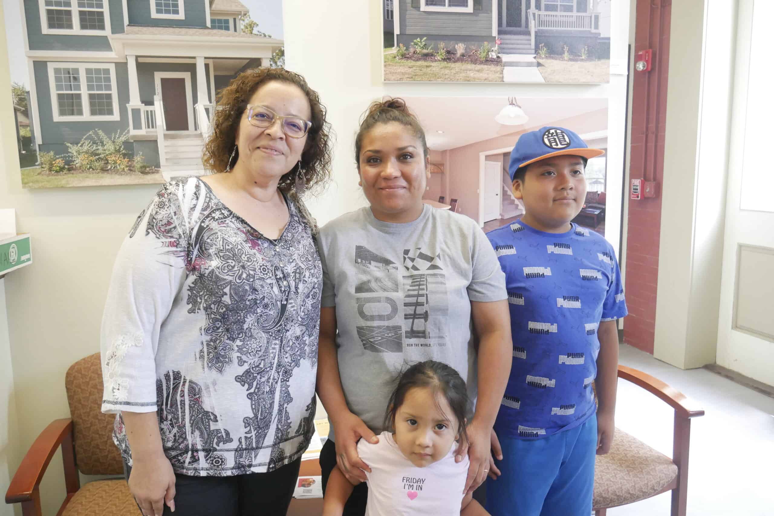 Rita and her family stand with Gloria, one of CHWC's bilingual housing counselors, in front of a photo of beautiful new homes.