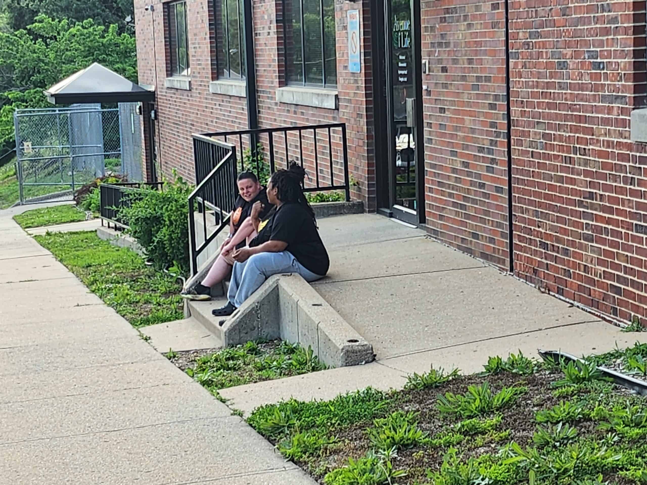 Two people sit outside on stairs outside a brick building, talking to each other.
